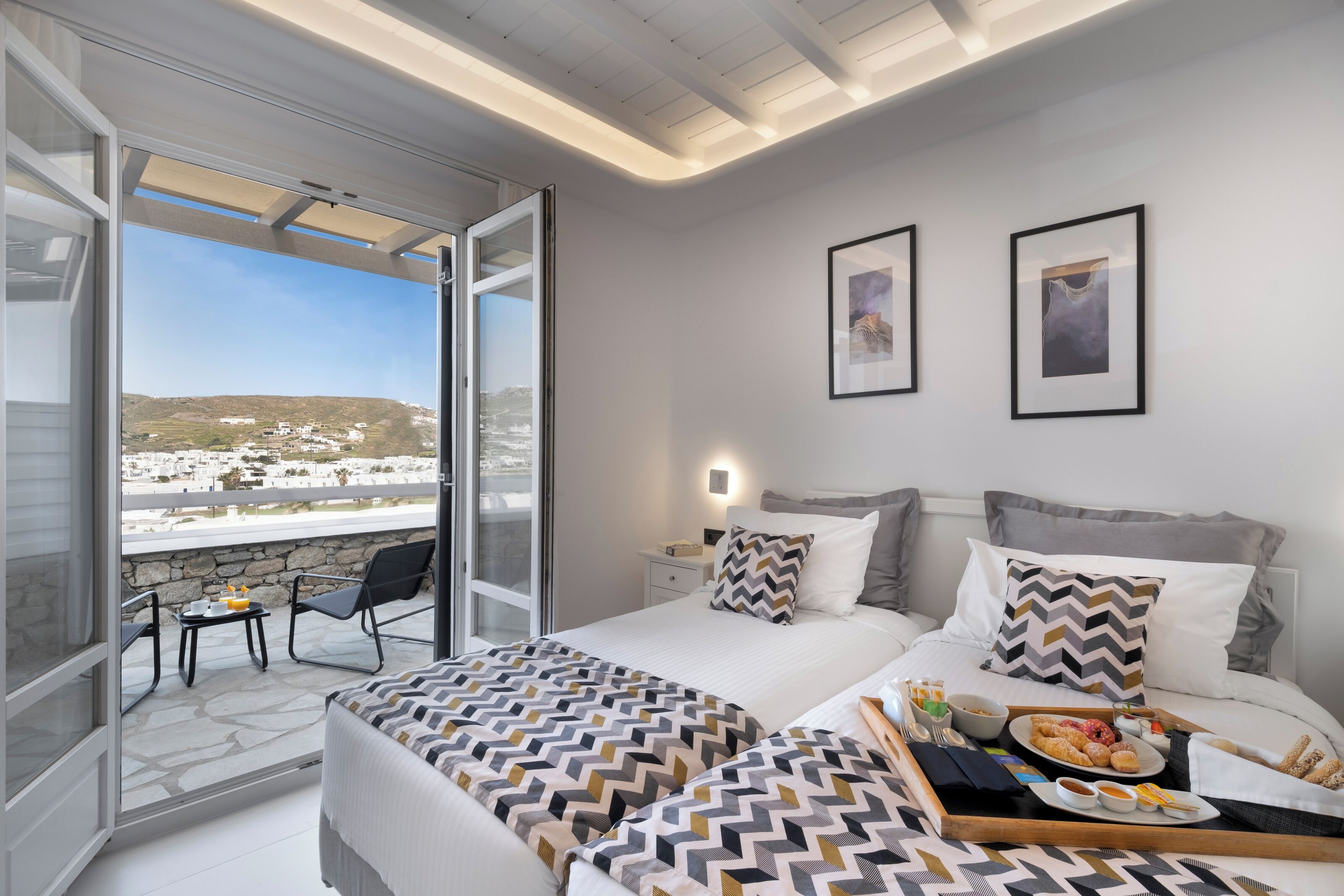 A hotel room with a balcony, bed with patterned bedding, a dining area with breakfast, and two framed pictures on the wall.