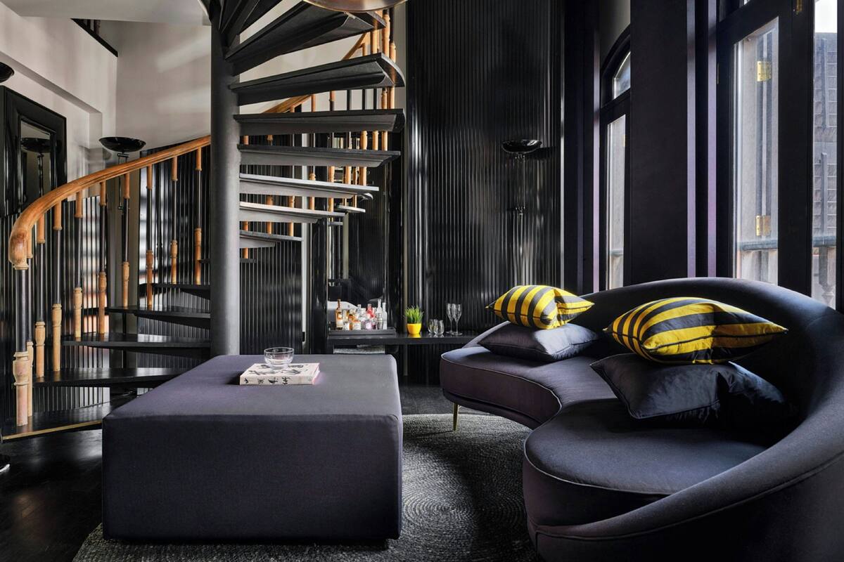 Un salon moderne aux couleurs sombres, avec un canapé noir orné de coussins à rayures, un pouf noir et un escalier en colimaçon en arrière-plan.