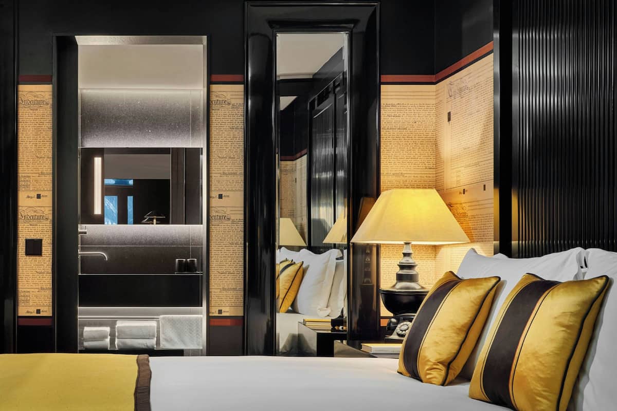 Une chambre d’hôtel avec un lit, une table de chevet, une lampe, un miroir et une salle de bain visible à travers une porte ouverte.