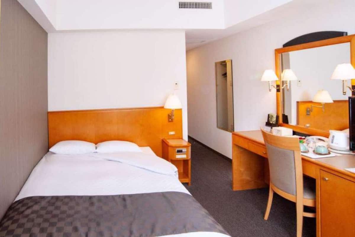 Una habitación de hotel con una cama, un escritorio, una silla, un espejo y una mesita de noche.