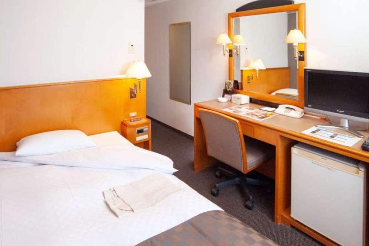 Una habitación de hotel con una cama, un escritorio con ordenador, un refrigerador pequeño y un espejo.