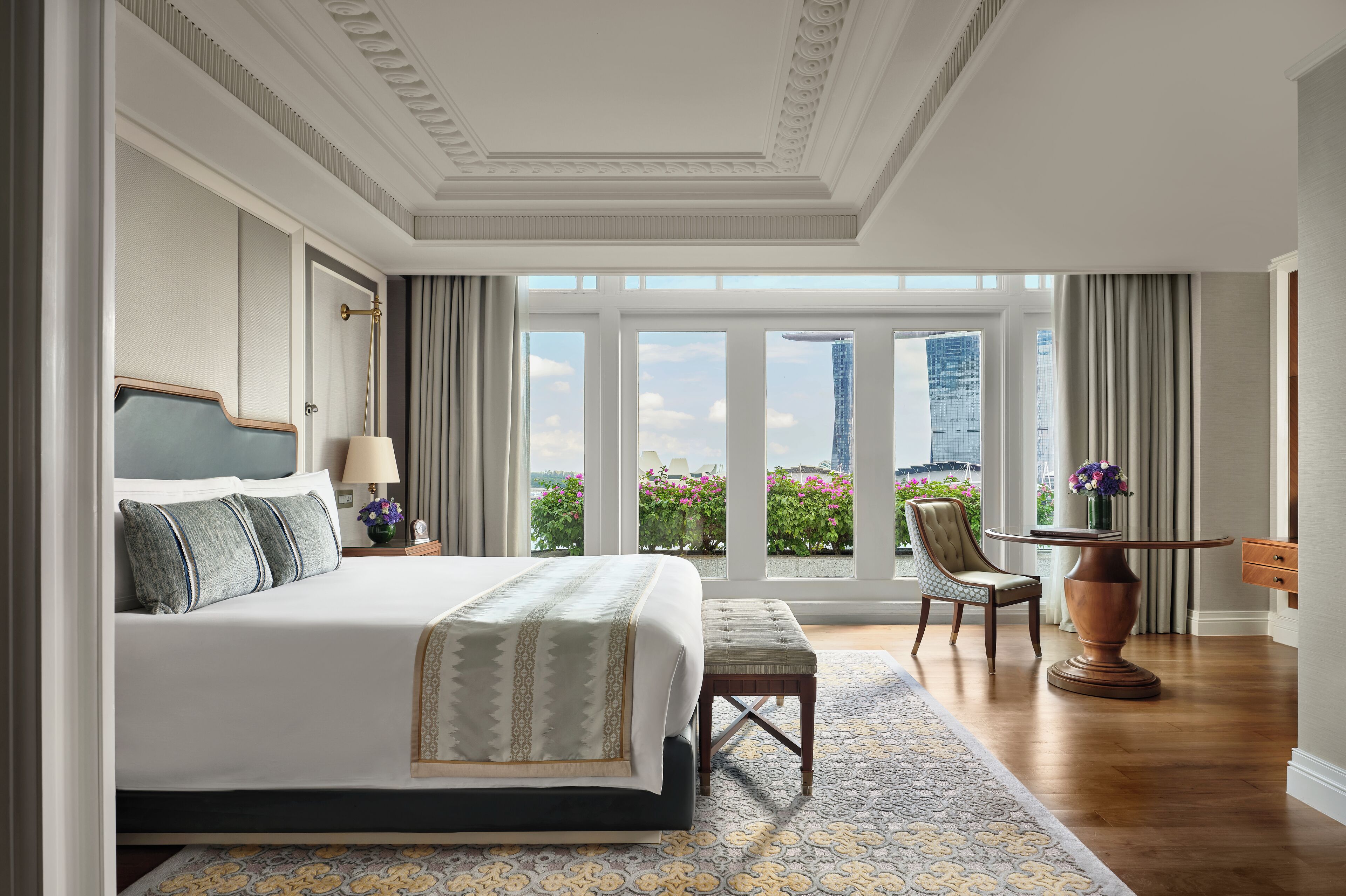 A spacious bedroom with a large bed, a sitting area with a table and chair, and a view of a cityscape through large windows.