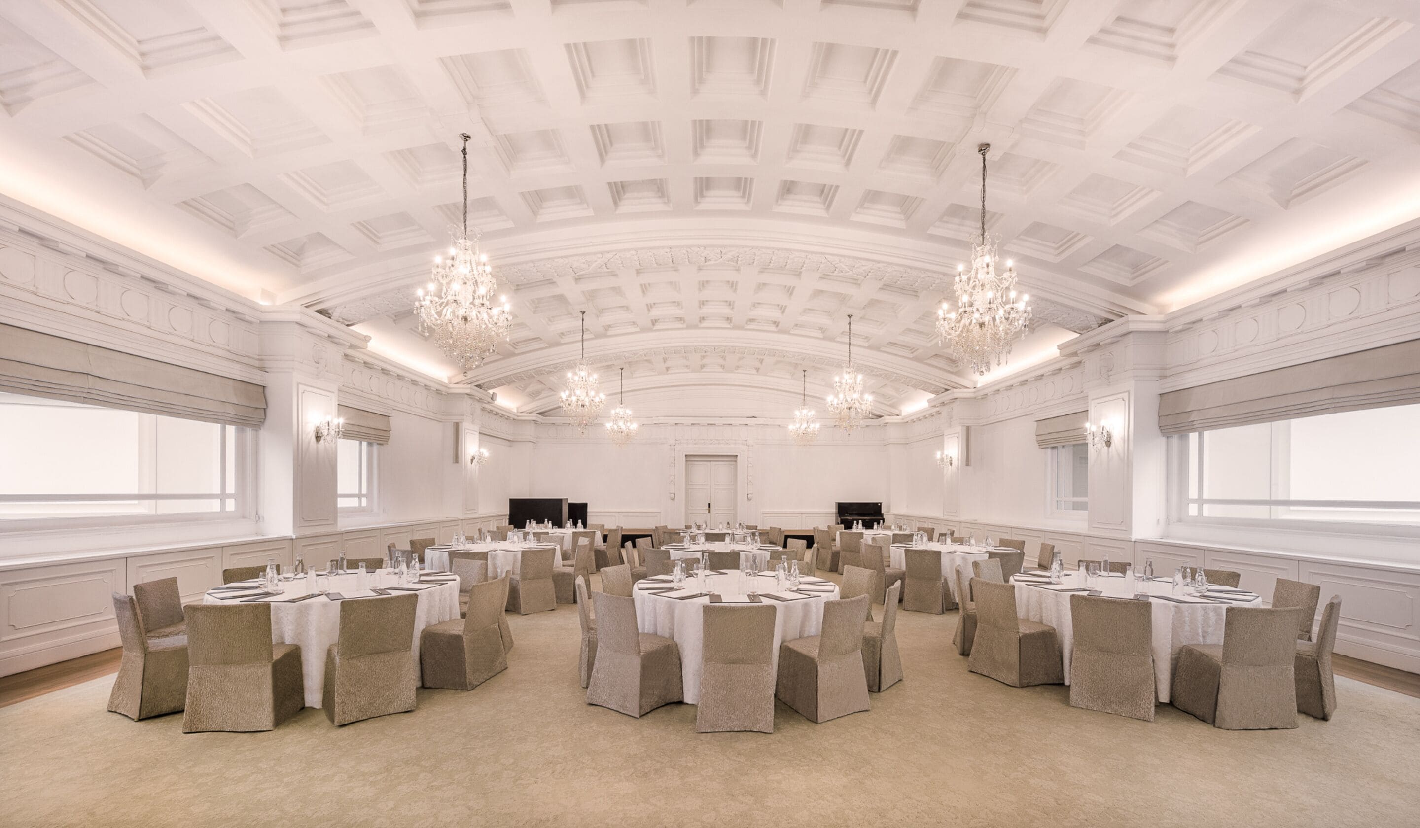 Un salón de baile imponente con techo ornamentado y candelabros, mesas redondas con manteles blancos y sillas color beige.
