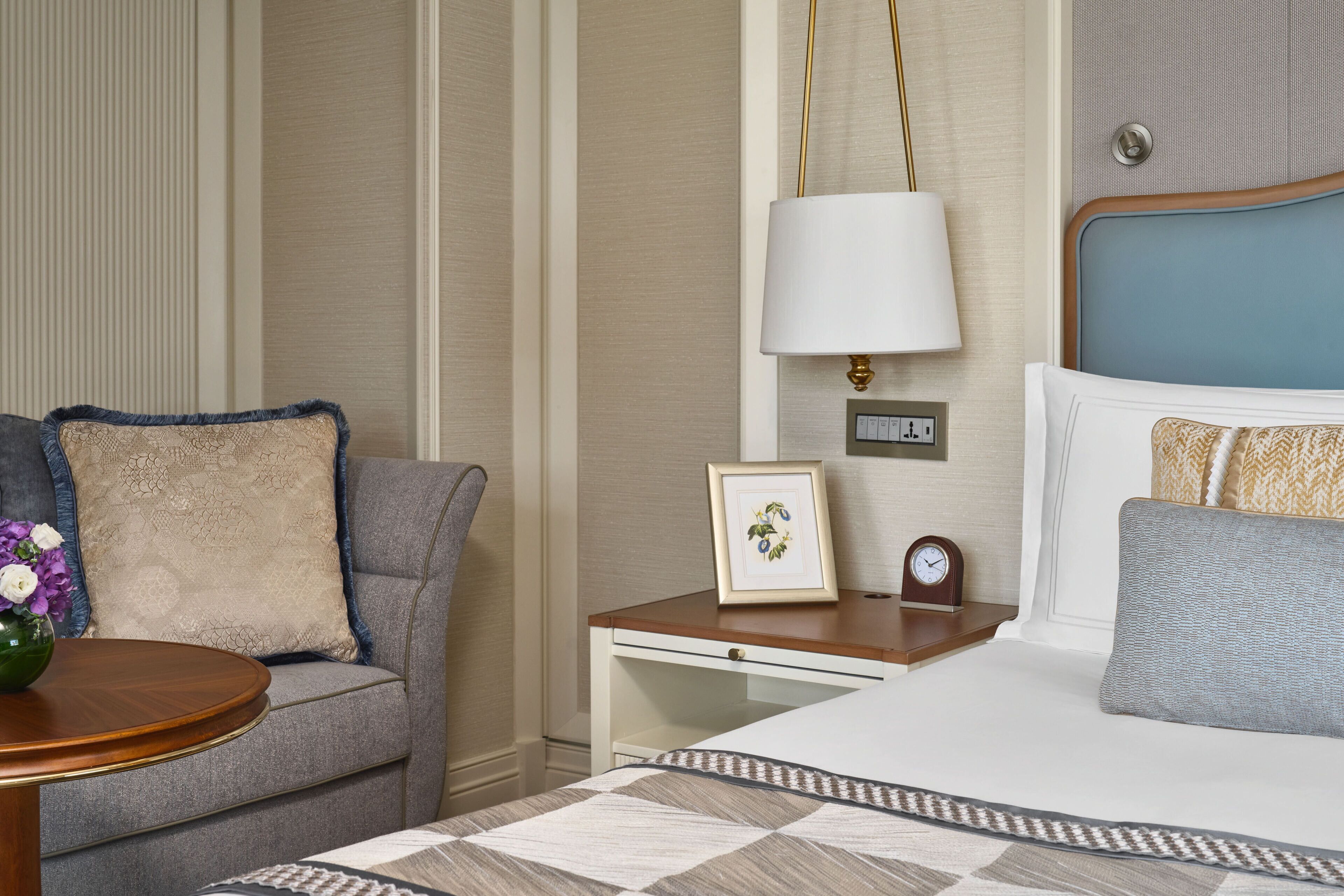 A hotel room with a bed, a bedside table with a lamp, a clock, and a framed picture.