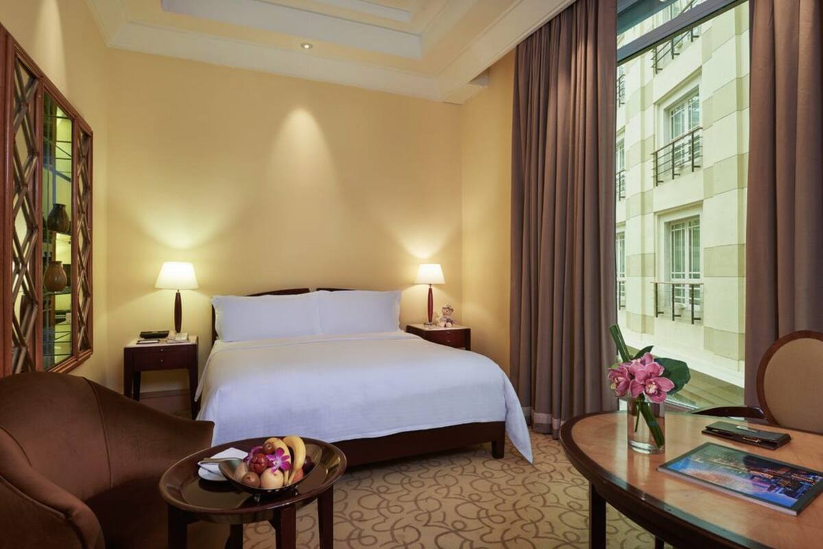 A hotel room with a bed, two bedside lamps, a small table with a bowl of fruit, a chair, and a view of a building through a window.