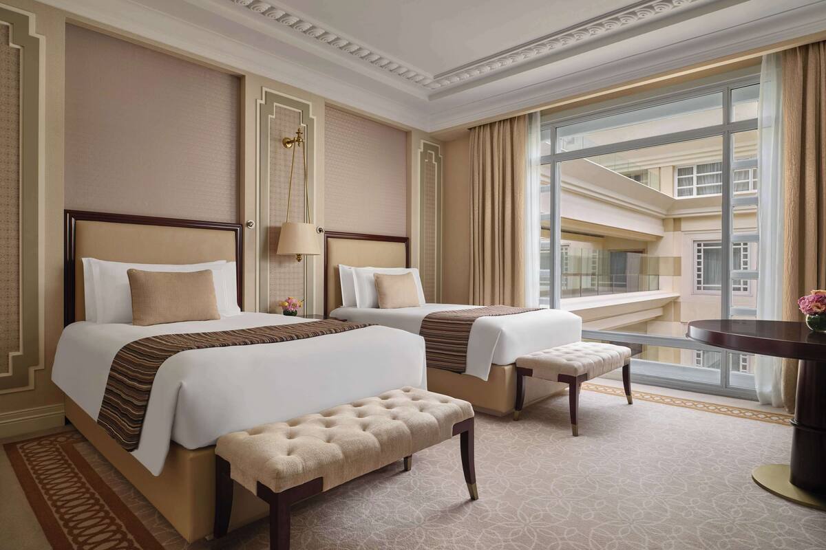A hotel room with two beds, a bench, a desk, and a large window offering a view of the building's interior.