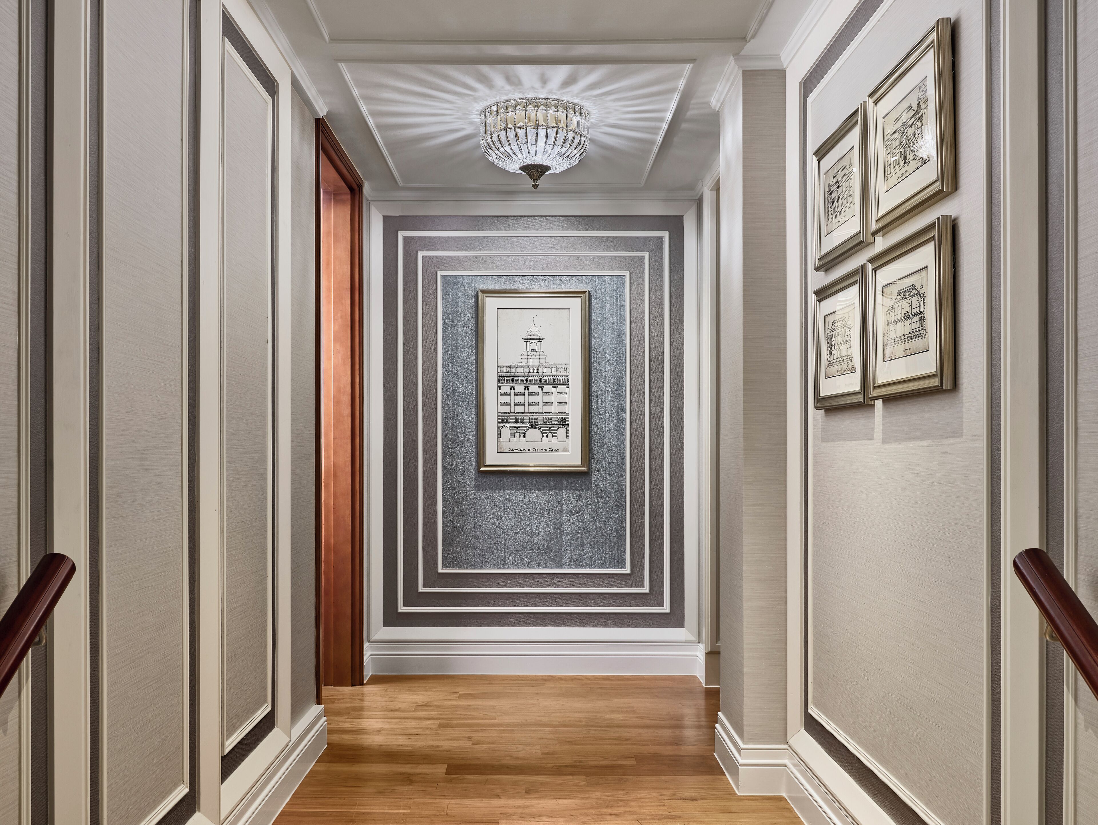 A hallway with a wooden floor, white walls with decorative molding, a chandelier, and framed architectural drawings on the wall.