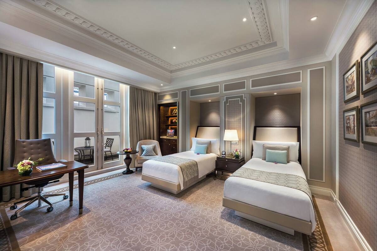 A hotel room with two beds, a desk, a chair, a small table with a lamp, and a bookshelf.