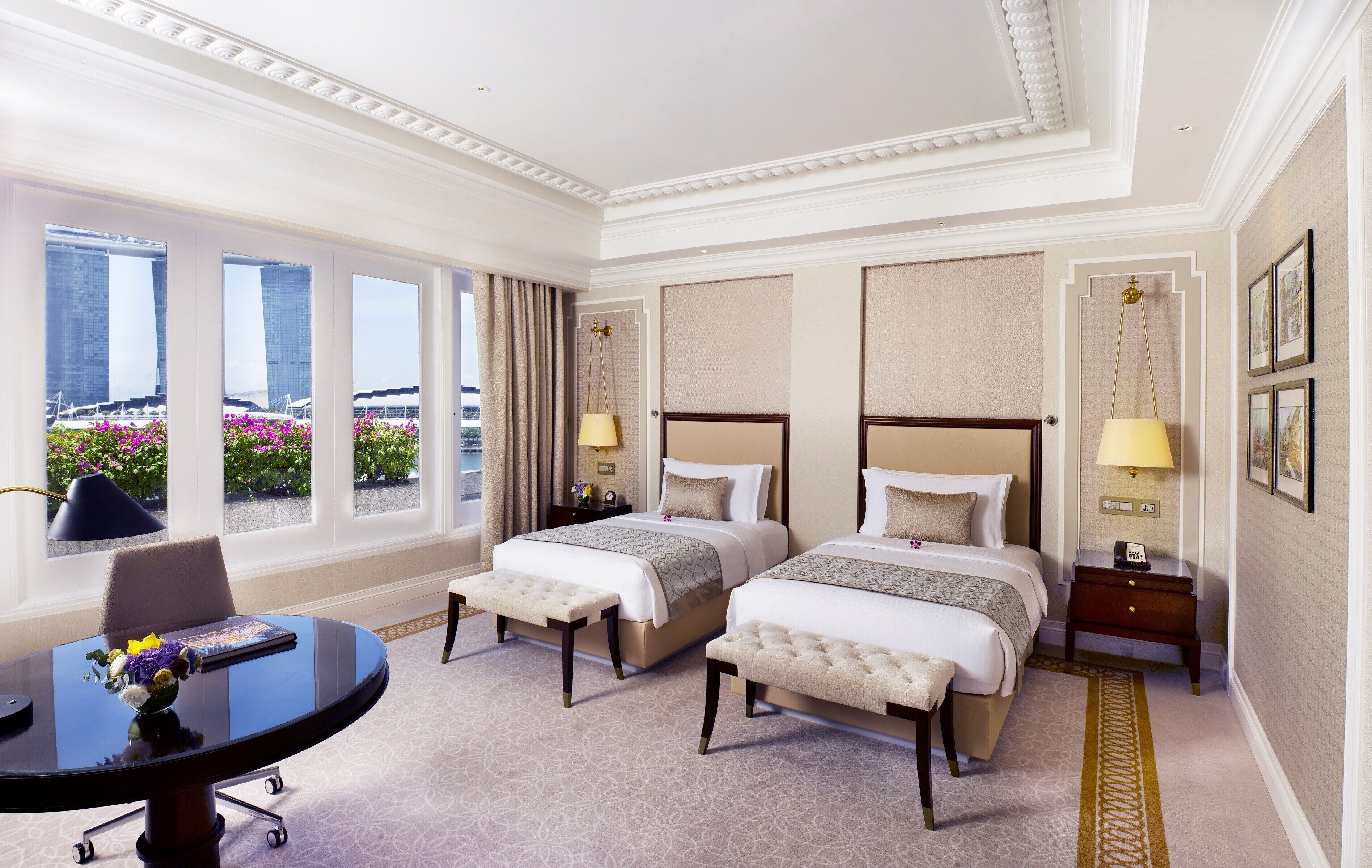 A hotel room with two beds, a desk, and a view of a building and flowers.