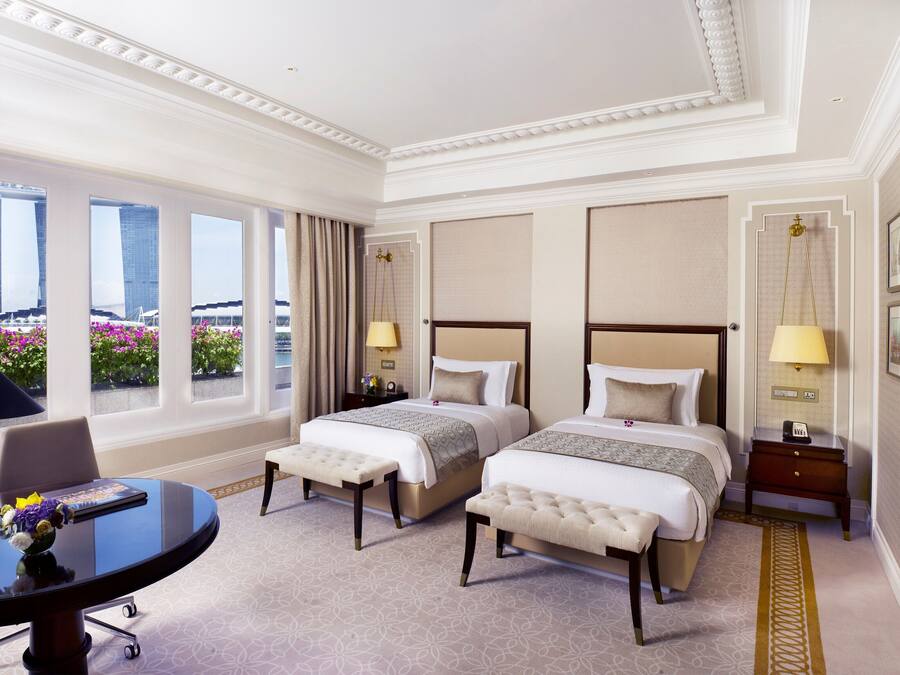 A hotel room with two beds, a desk, and a view of a building and flowers.