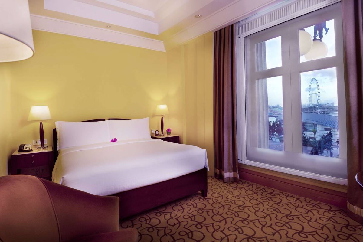 A hotel room with a large bed, two bedside lamps, a nightstand, a chair, and a large window with a view of a Ferris wheel.