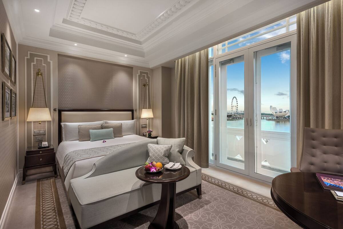 A hotel room with a large bed, a sofa, a small table with fruit, and a view of a Ferris wheel through a large window.