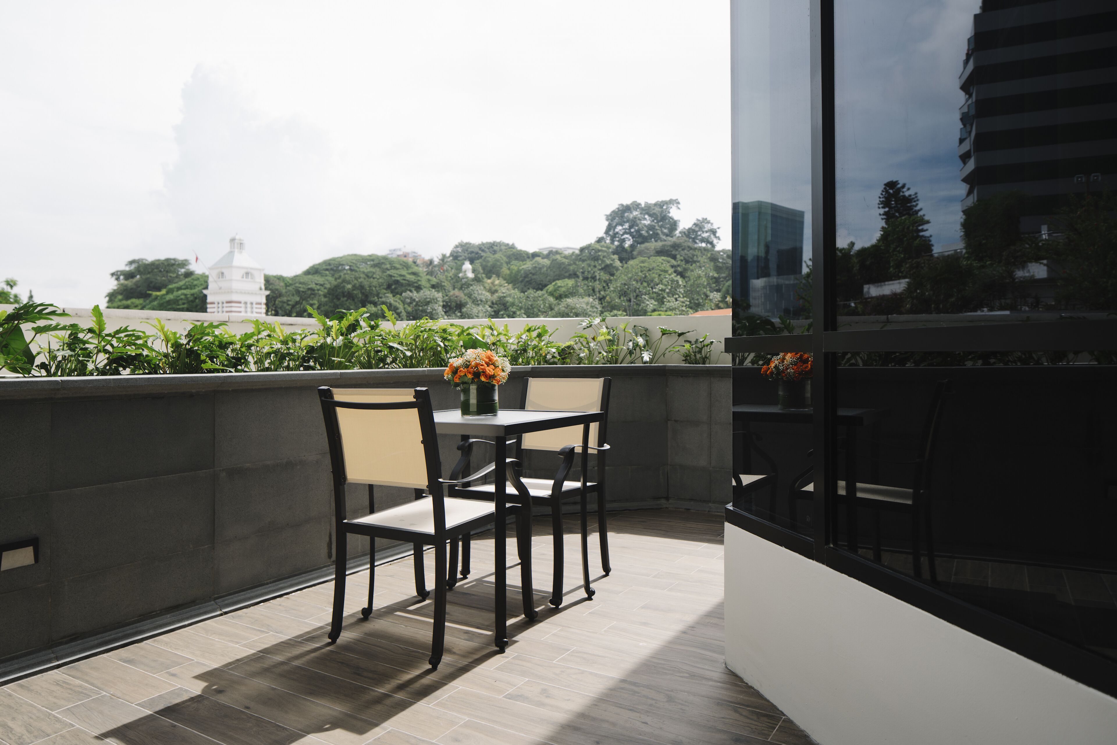 A balcony with a table and chairs, a vase of flowers, and a view of greenery and a white building in the background.