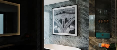 A modern bathroom with a marble wall, a framed picture of a bridge, a white bathtub, and a glass shelf with toiletries.