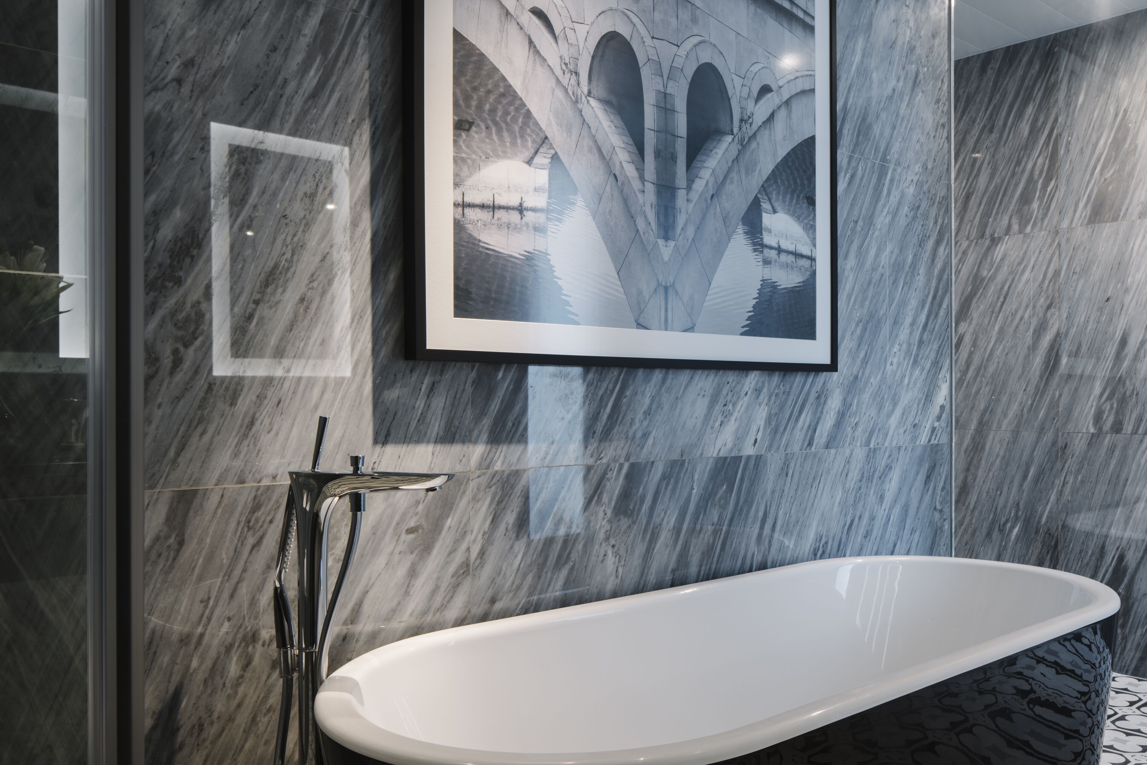 Une salle de bain moderne avec un mur en marbre, un tableau encadré représentant un pont et une baignoire sur pieds.