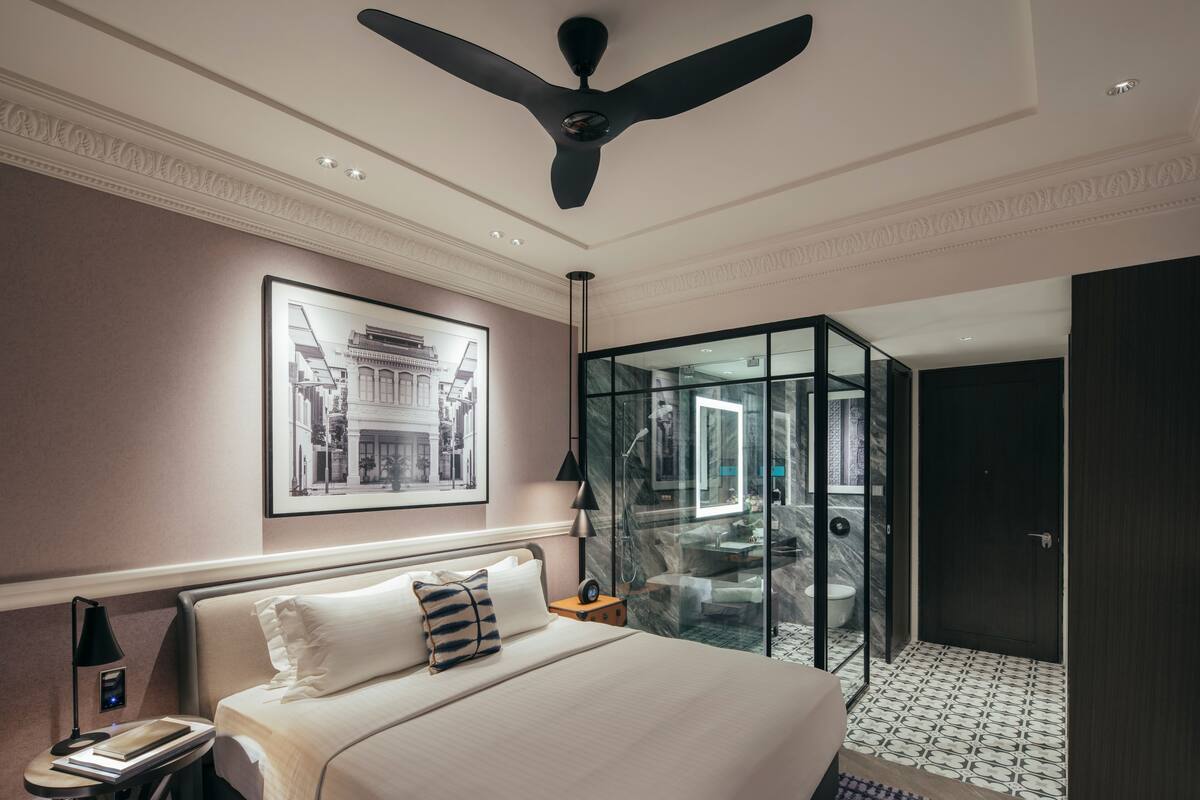 A hotel room with a bed, a ceiling fan, a framed picture on the wall, and a glass-enclosed bathroom.