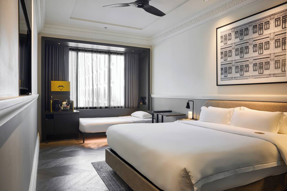 A hotel room with a large bed, a smaller bed, a desk, a chair, a ceiling fan, a window with curtains, and a framed picture on the wall.