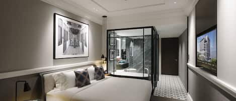A modern hotel room with a large bed, a glass-enclosed bathroom, and a view of a cityscape.