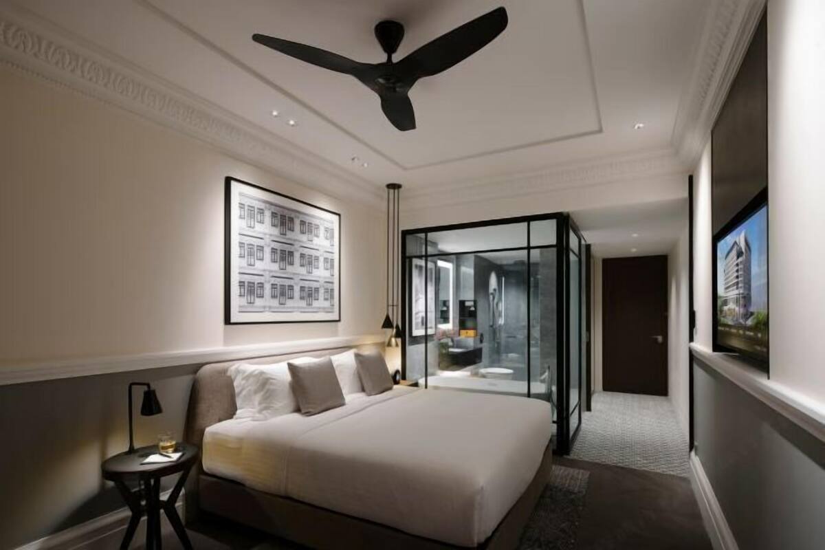 A hotel room with a bed, a ceiling fan, a glass partition, and a framed picture on the wall.