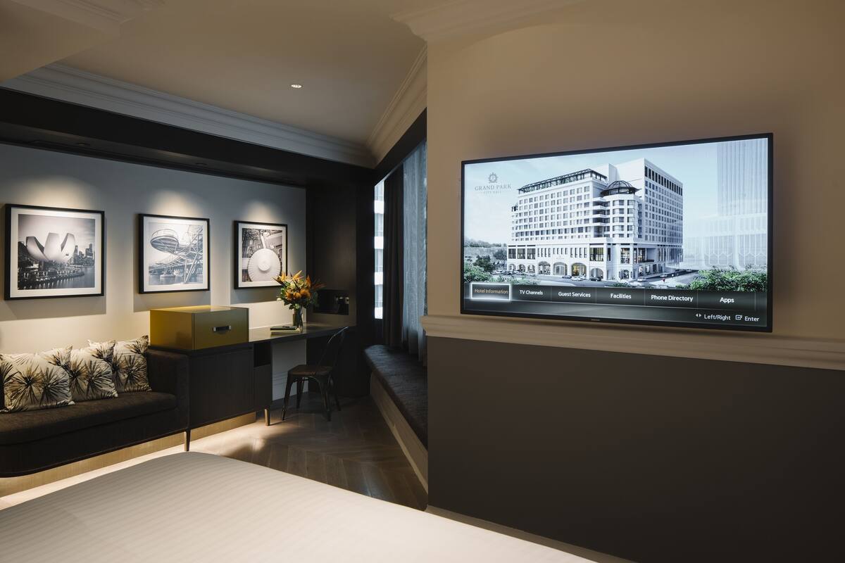 A modern hotel lobby with a large TV displaying a building image, framed artwork on the wall, a sofa, and a desk with a vase of flowers.