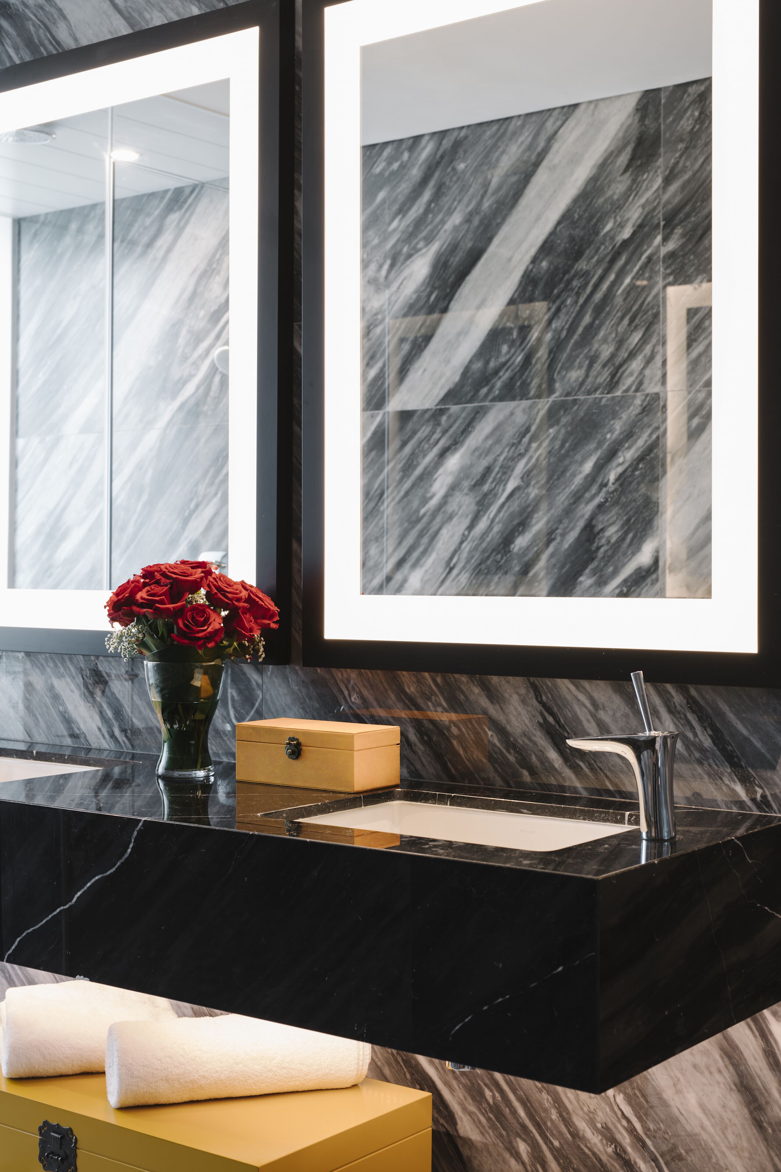 A modern bathroom with a marble backsplash, a black countertop, a sink with a chrome faucet, a vase with red roses, and a mirror.