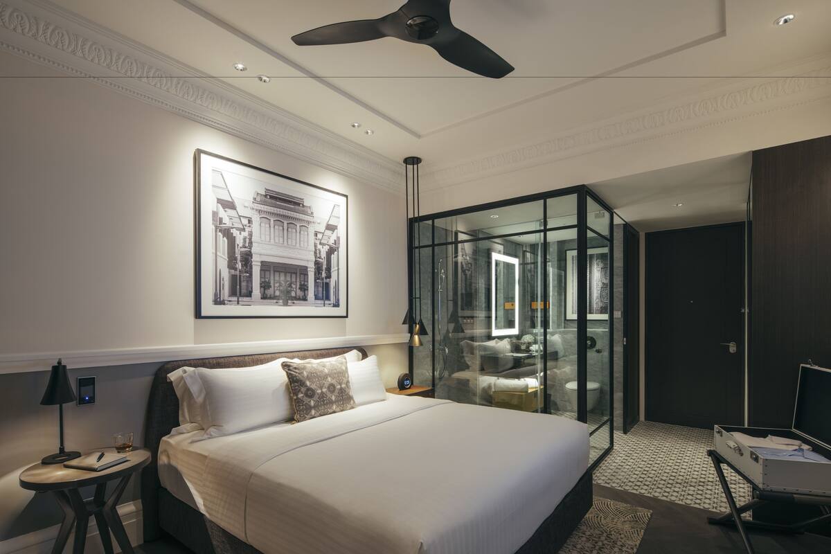 A modern hotel room with a large bed, a glass partition, a ceiling fan, and a framed picture on the wall.