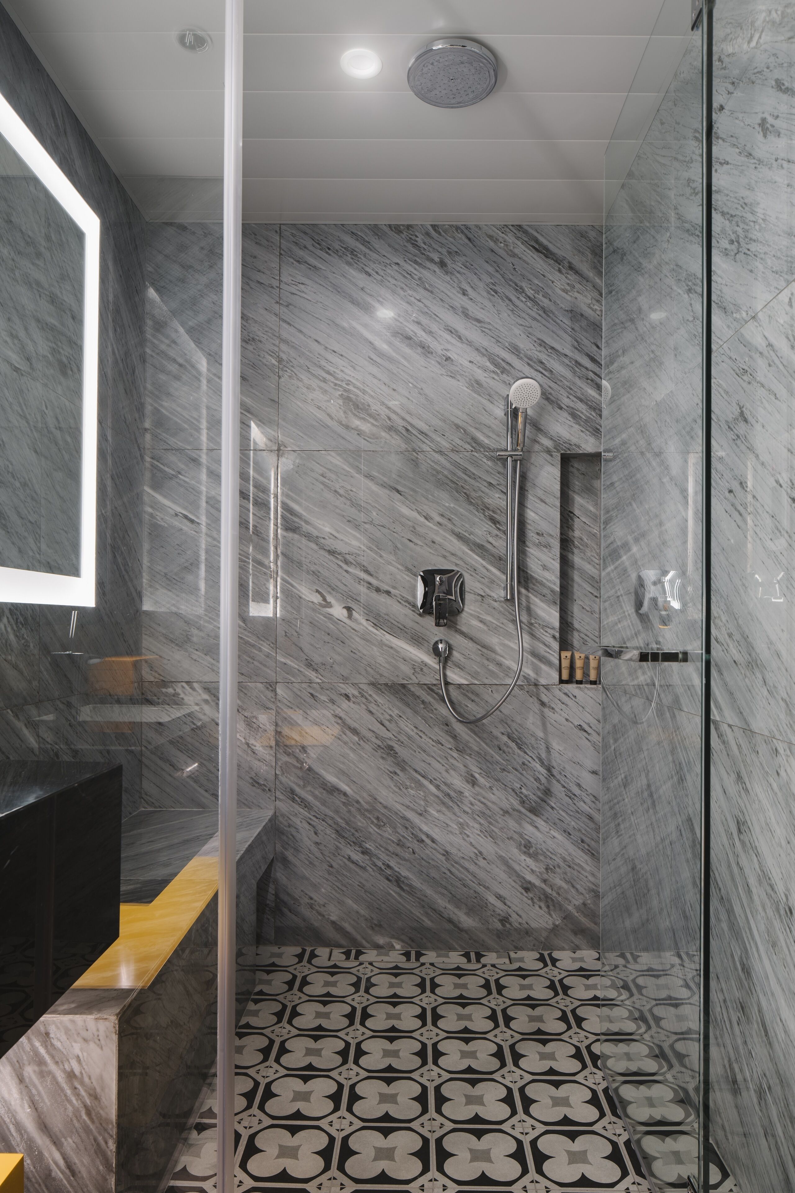 A modern bathroom with a glass shower enclosure, marble walls, and patterned floor tiles.