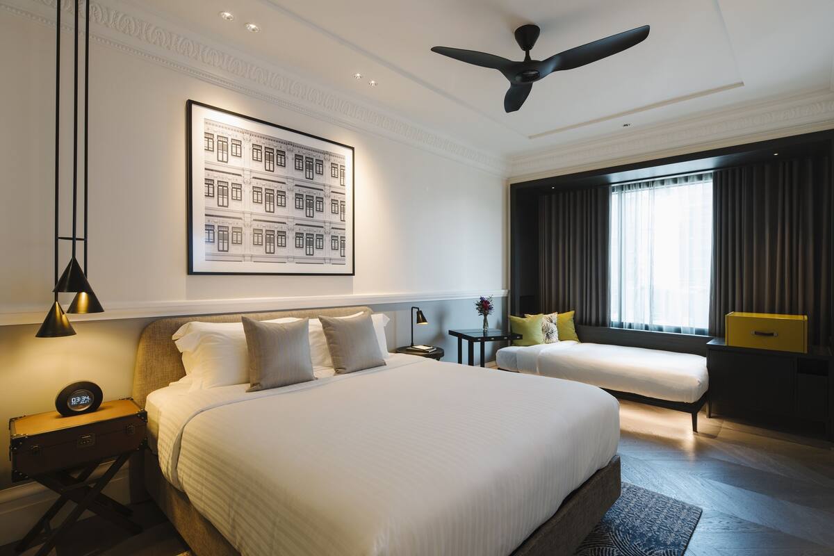 A modern hotel room with a large bed, a smaller bed, a ceiling fan, a framed cityscape artwork, and a window with curtains.