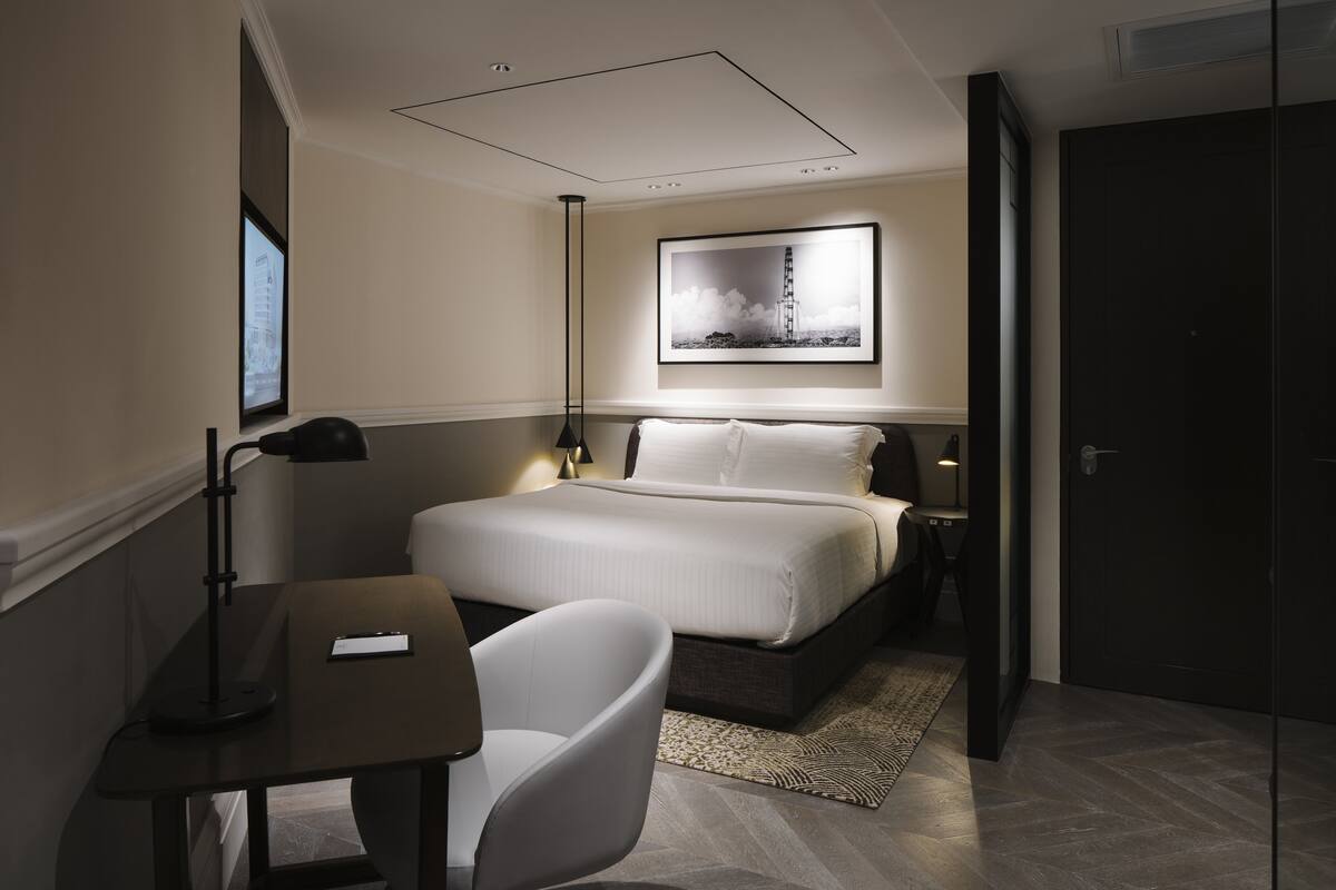 A modern hotel room with a neatly made bed, a desk with a chair, a large framed picture on the wall, and a dark door.