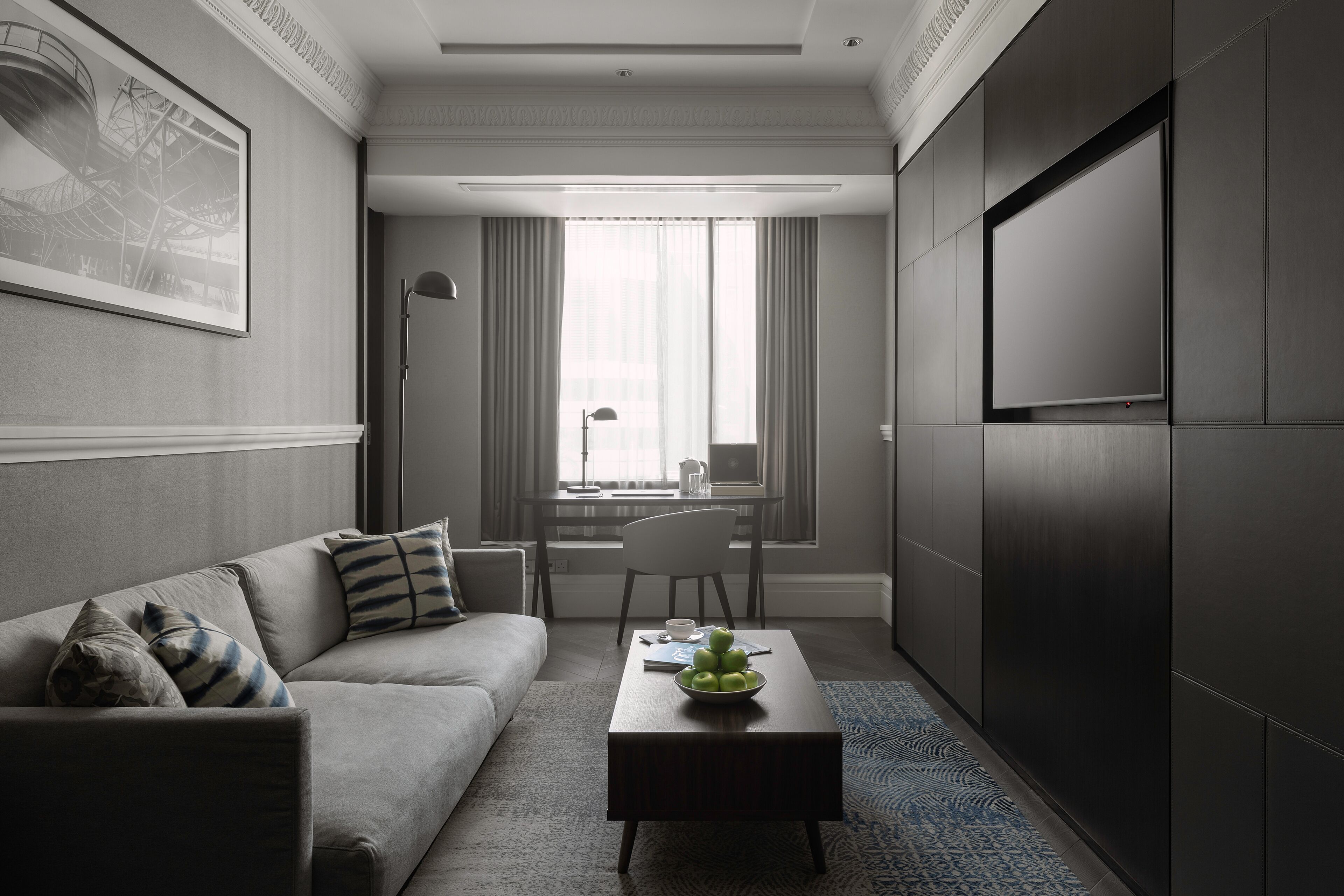 A modern hotel room with a sofa, a coffee table with a fruit bowl, a desk, and a large TV screen.