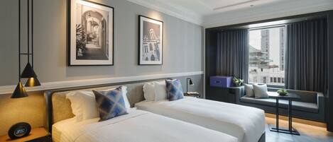 A hotel room with two beds, a ceiling fan, a nightstand with a clock, and a window with a view of a building.