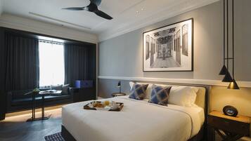 Une chambre d’hôtel moderne avec un grand lit, un bureau avec une chaise, une fenêtre avec des rideaux, un ventilateur de plafond et un tableau encadré au mur.
