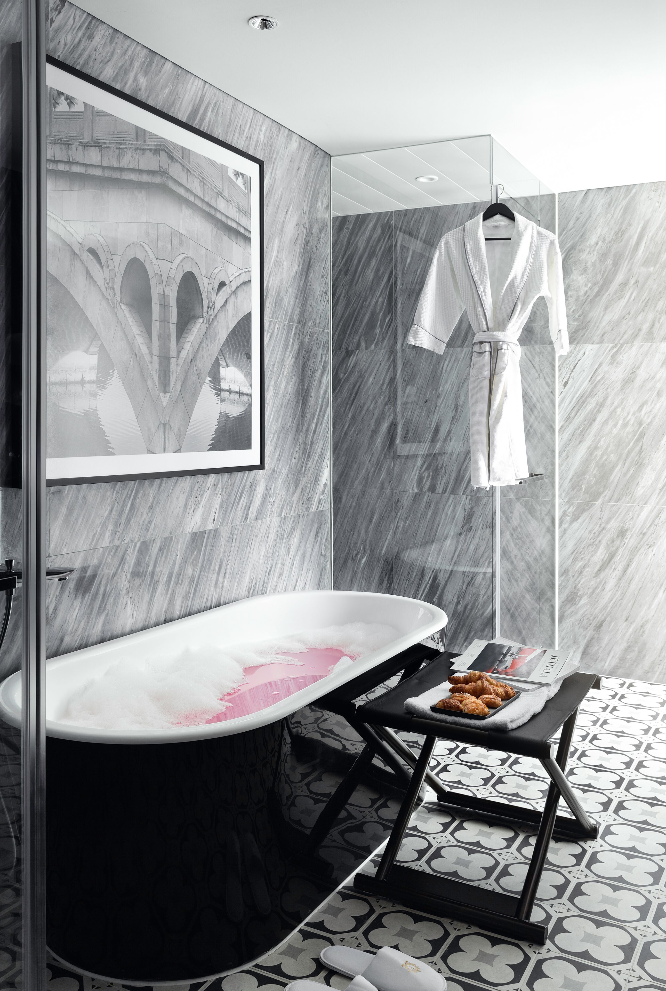 A modern bathroom with a freestanding bathtub, a framed picture on the wall, a robe hanging on a rack, and a tray with croissants on a small table.