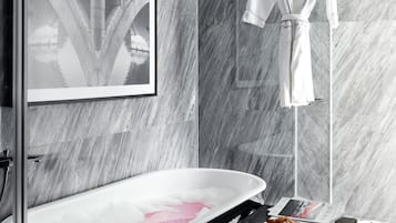 A modern bathroom with a freestanding bathtub, a framed picture on the wall, a robe hanging on a rack, and a tray with croissants on a small table.