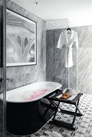 A modern bathroom with a freestanding bathtub, a framed picture on the wall, a robe hanging on a rack, and a tray with croissants on a small table.