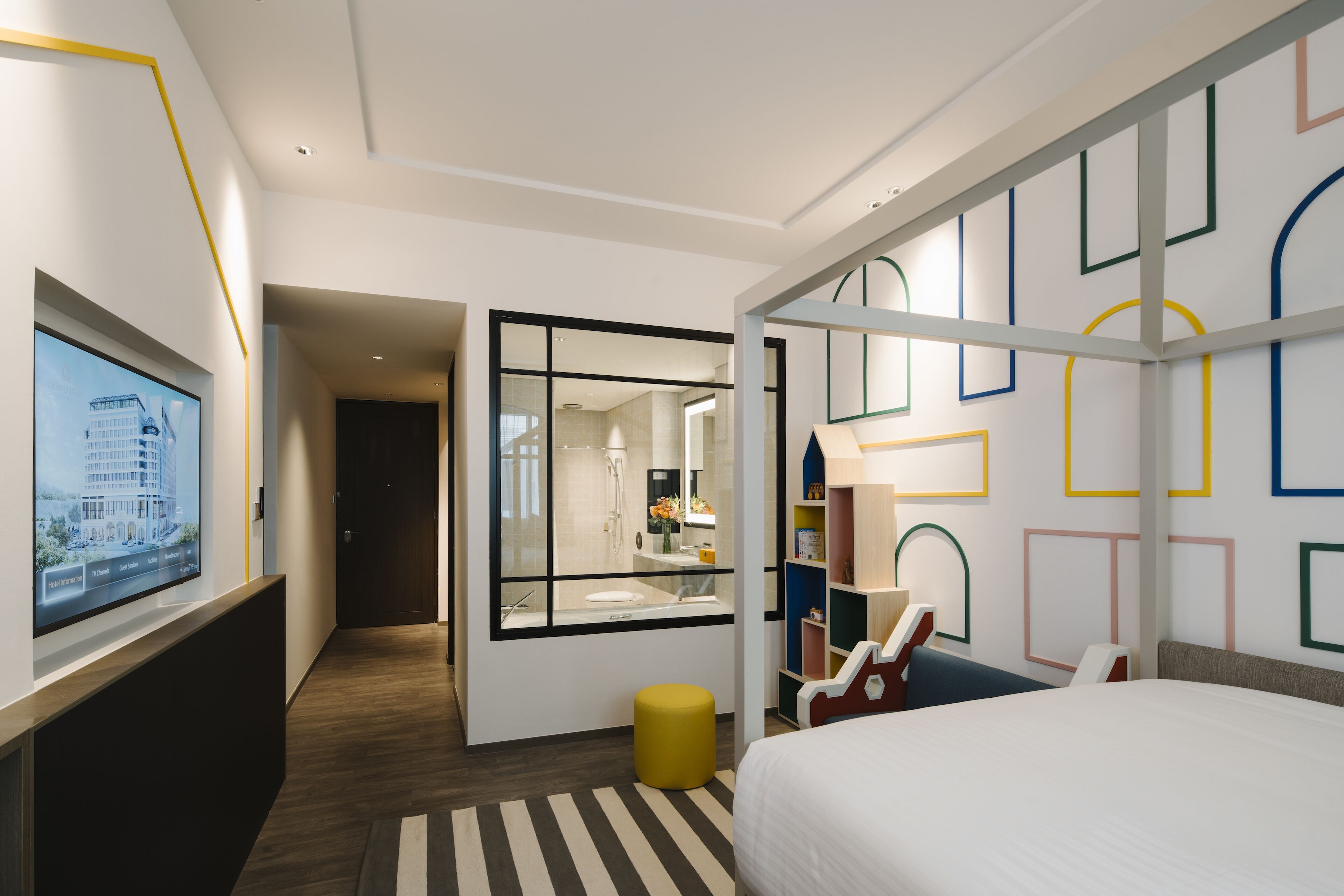 A modern hotel room with a large bed, a television, a bathroom with a mirror, and a hallway leading to another room.