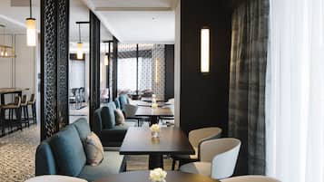 A modern hotel lobby with a central seating area, a bar, and a view of the cityscape through large windows.