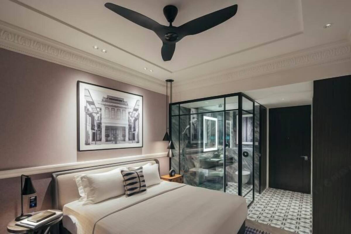 A hotel room with a bed, a ceiling fan, a framed picture on the wall, and a glass-enclosed bathroom.