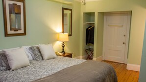 A neatly made bed with a patterned bedspread and pillows, a bedside lamp, a coat rack with a jacket, and a door to a closet. - Beacon Inn 1750 (Brookline)