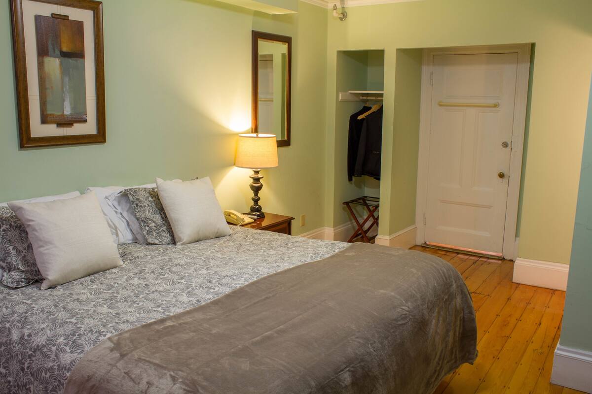 Una cama bien hecha con colcha y almohadas estampadas, una lámpara de noche, un perchero con una chaqueta y la puerta de un armario.
