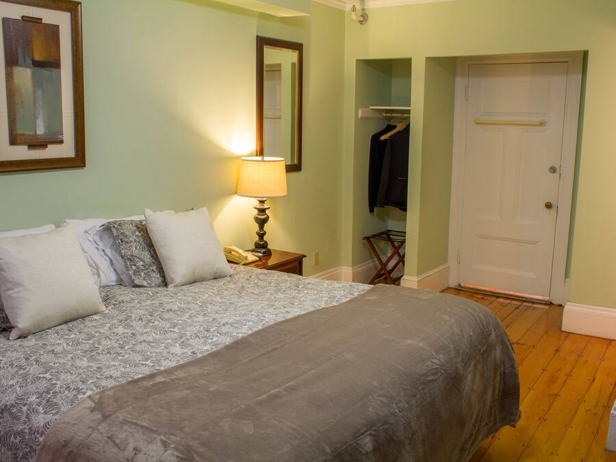 A neatly made bed with a patterned bedspread and pillows, a bedside lamp, a coat rack with a jacket, and a door to a closet.