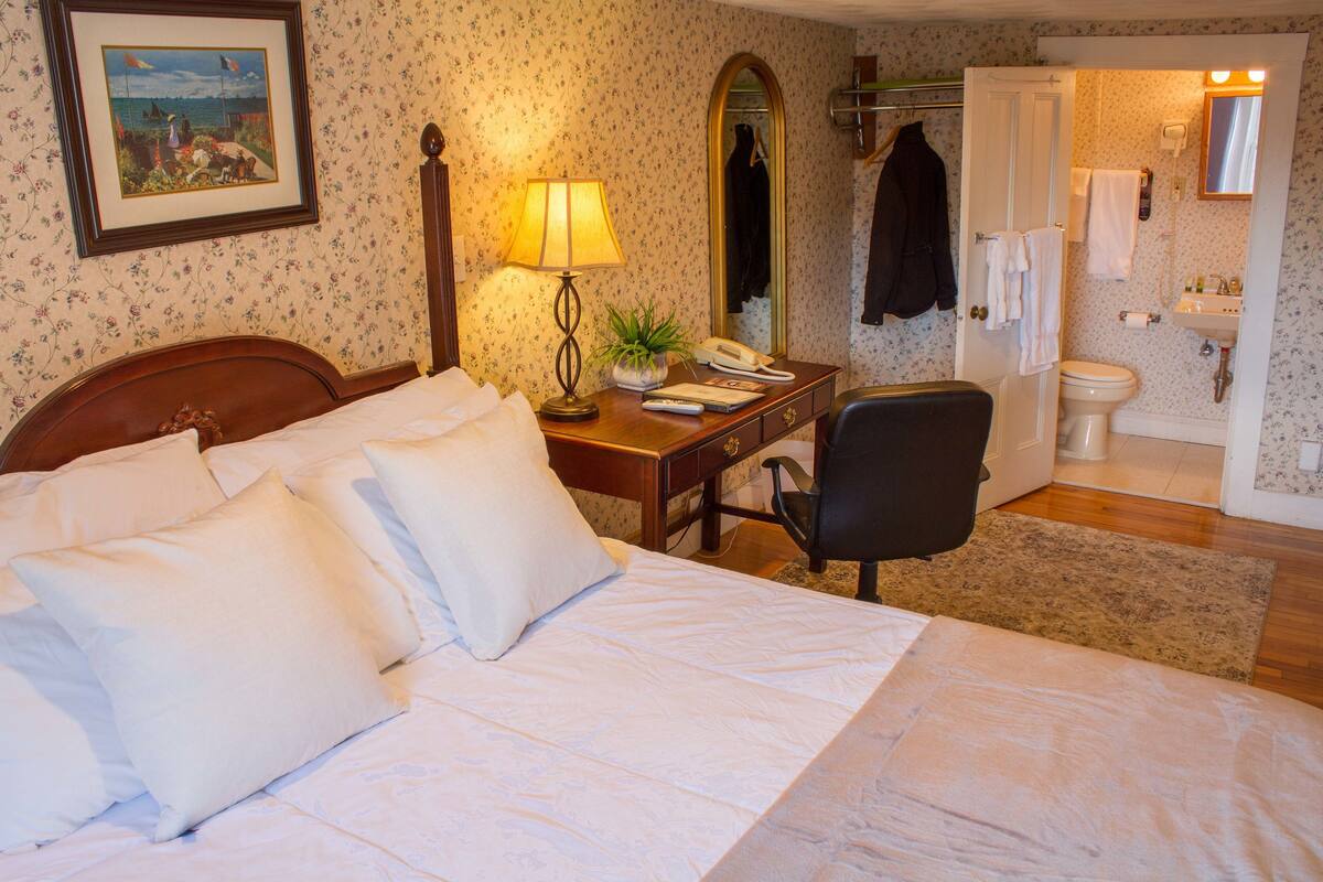 Habitación de hotel con cama, escritorio con lámpara, silla, espejo y baño con inodoro y lavamanos.