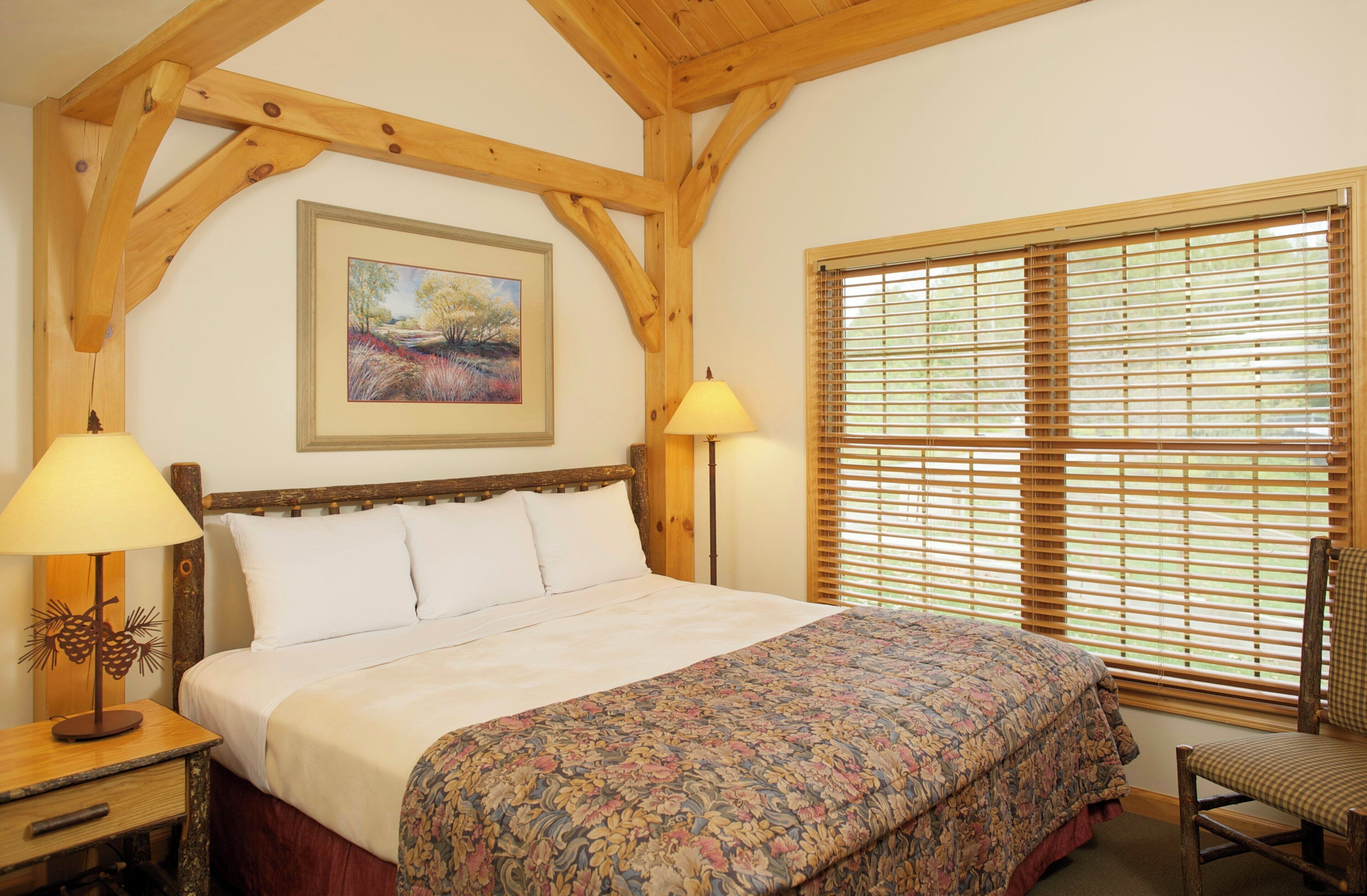 A bedroom with a wooden ceiling, a bed with white linens, a floral bedspread, a painting on the wall, and a window with blinds.