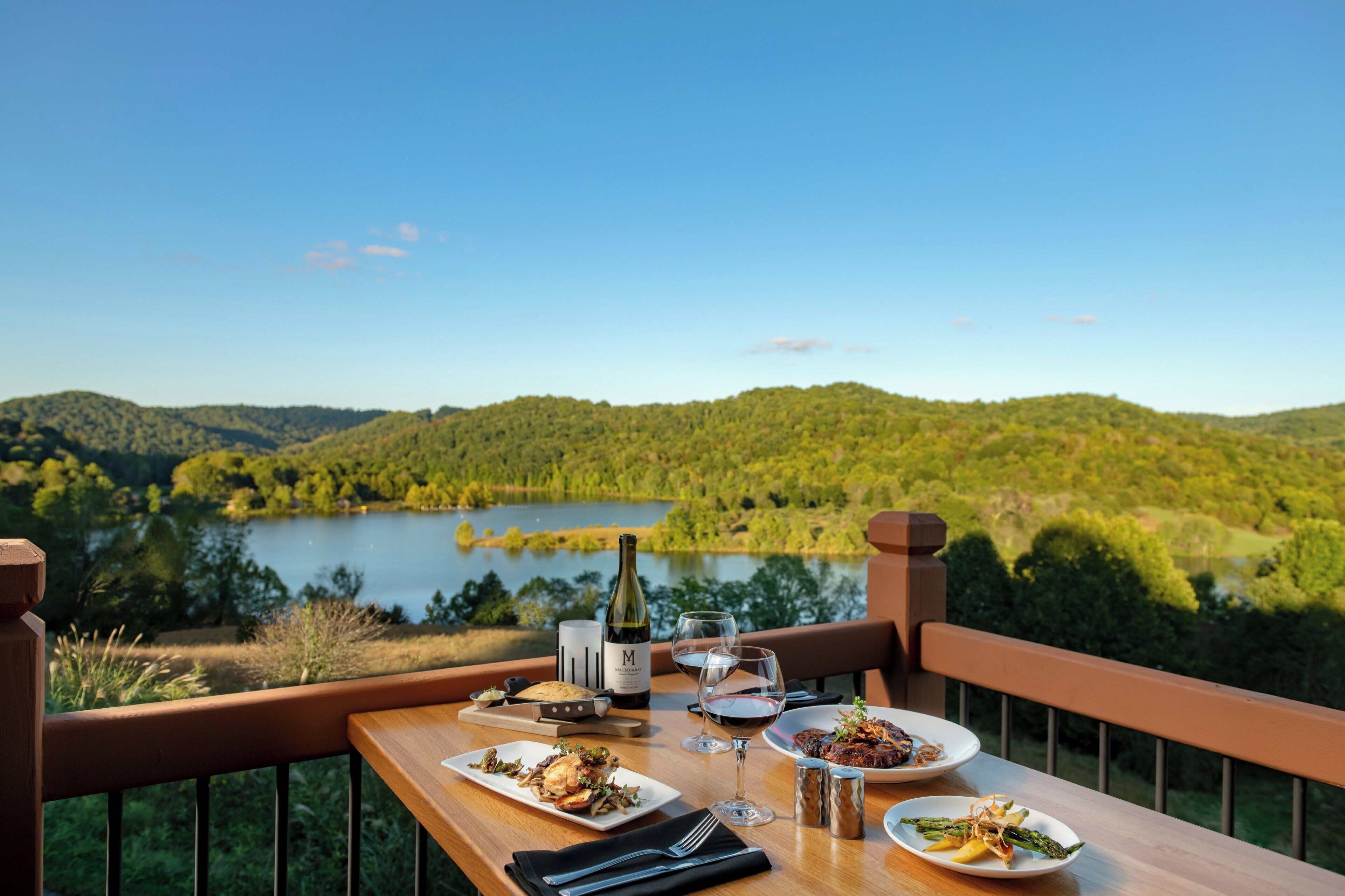 Una mesa de comedor con platos de comida, vasos y una botella, con vista a un lago y colinas boscosas.