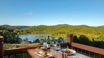 Una mesa de comedor con platos de comida, vasos y una botella, con vista a un lago y colinas boscosas.