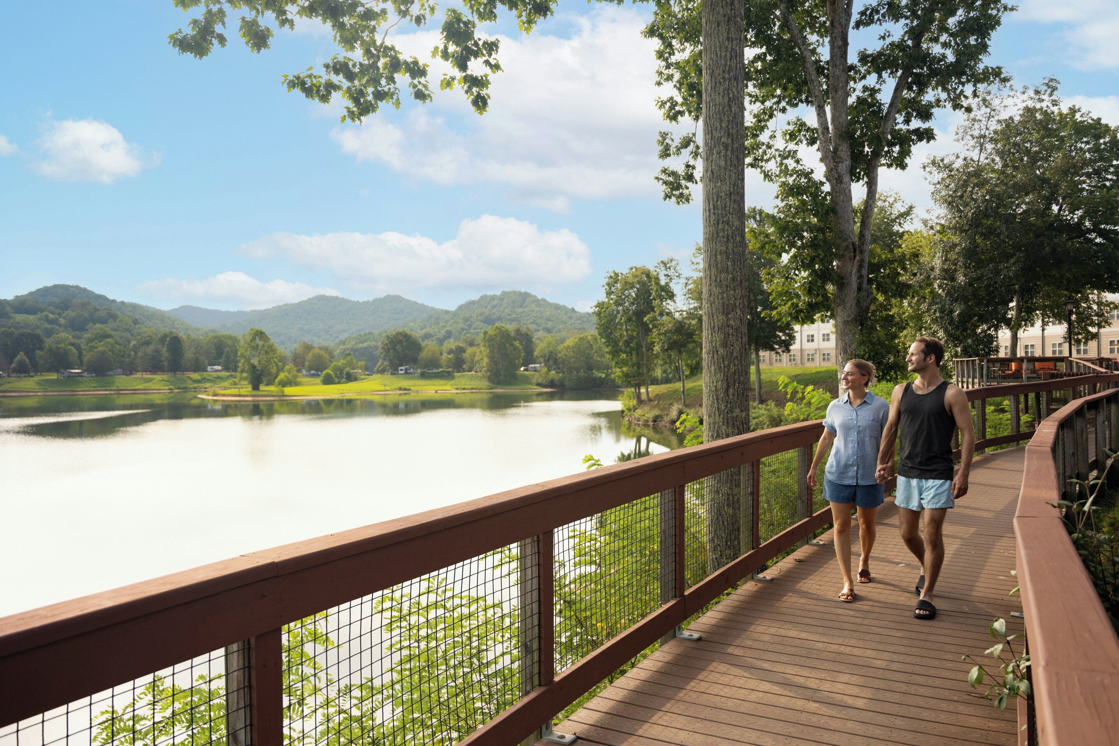 Una passerella in legno con ringhiera metallica, con vista su un lago calmo e colline verdi.