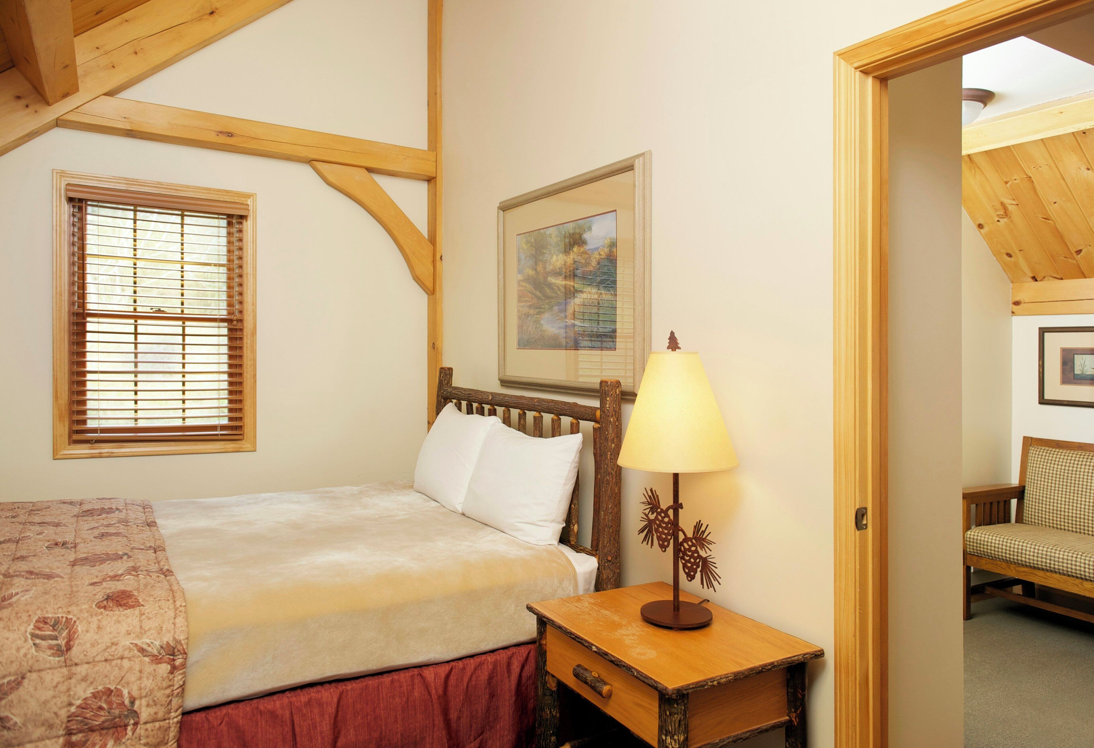 Un dormitorio con cama, mesita de noche, lámpara, ventana con persianas, un cuadro en la pared y una puerta que da a otra habitación.