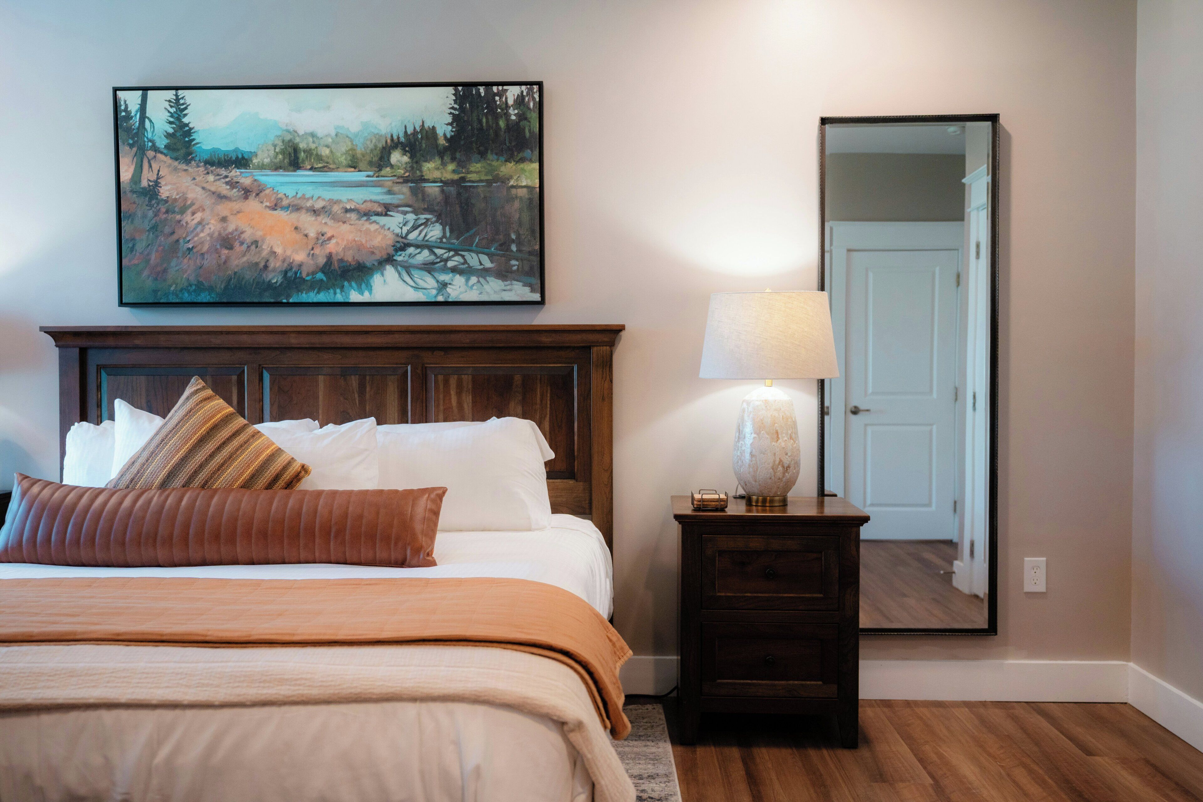 A bedroom with a bed, pillows, a nightstand with a lamp, a painting on the wall, and a door.