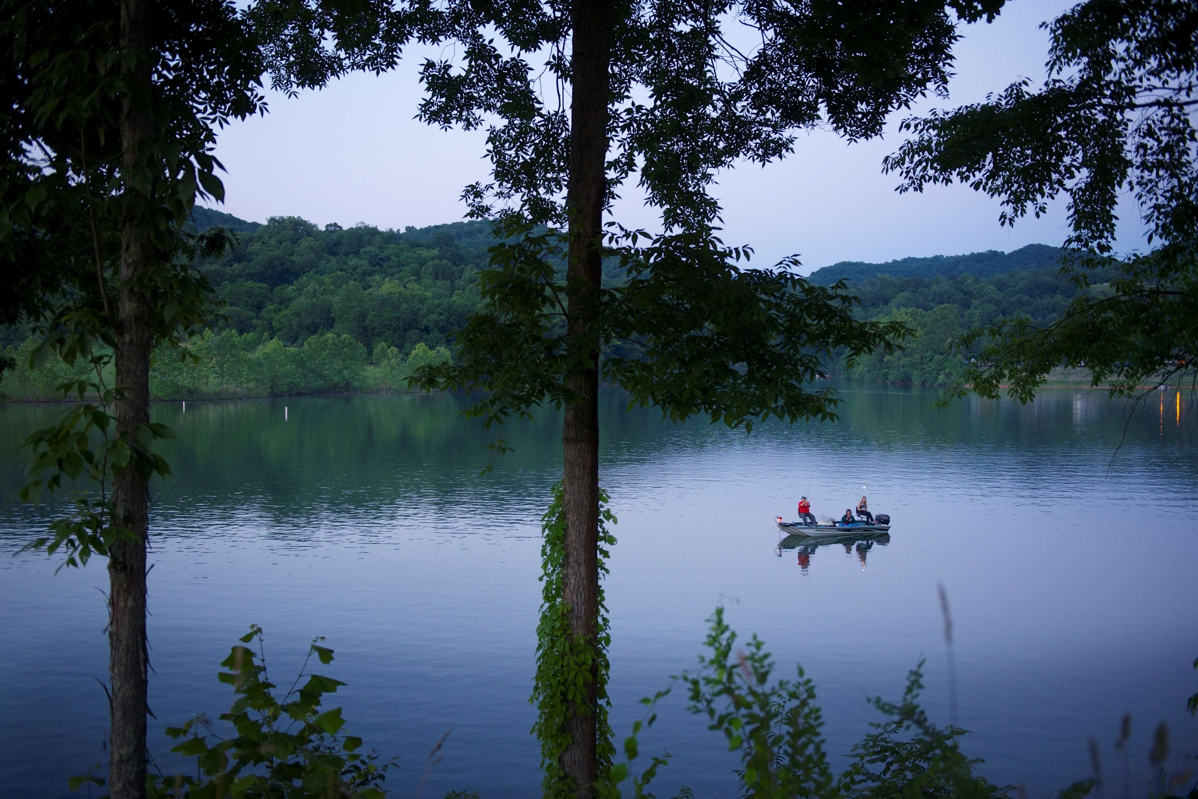 A calm lake with a boat carrying people, surrounded by trees and hills.