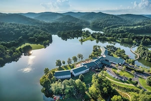 Un edificio grande con techo verde, rodeado por un lago y colinas boscosas.